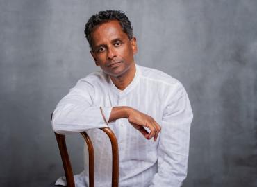 A photo of Shyam sitting backwards on a chair, looking straight into the camera. He wears a white blouse and the background is grey.