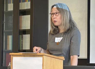 A photo of Professor Lee during her presentation. She lectures from behind a podium, her hand resting upon it. Behind her is a bookshelf and whiteboard. She looks toward the audience.
